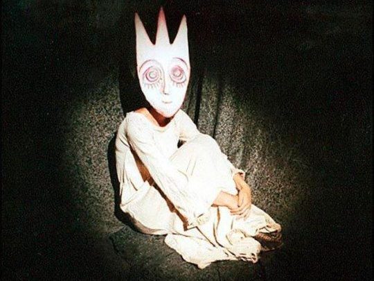 A figure in a white dress sits on the ground, wearing a large, stylized mask with a crown shape and big eyes, against a dark background.